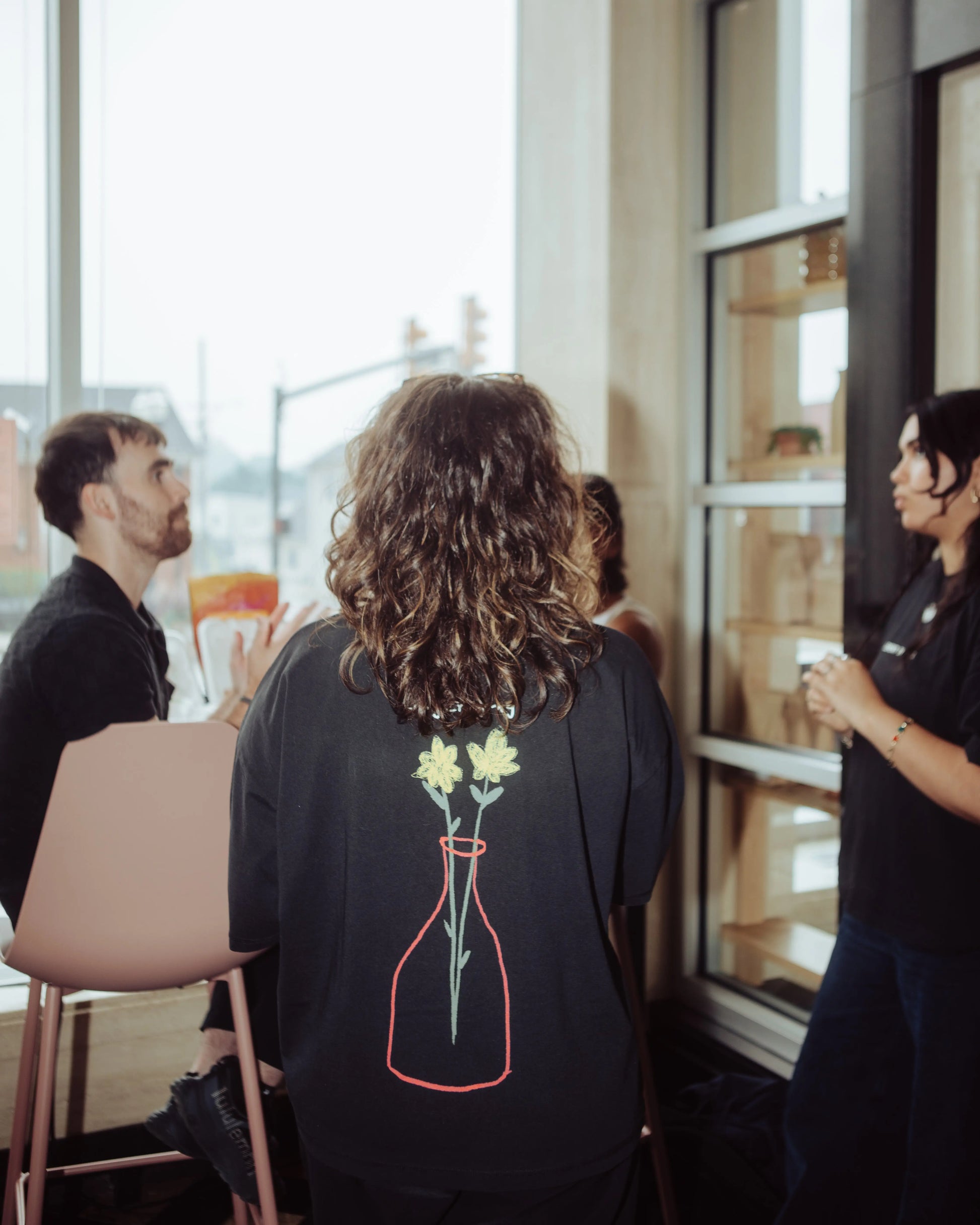 A person standing indoors with their back to the camera, wearing a black sweatshirt with a graphic of yellow flowers in a red-outlined vase on the back. The scene is set in a cafe or modern interior with other people and large windows.