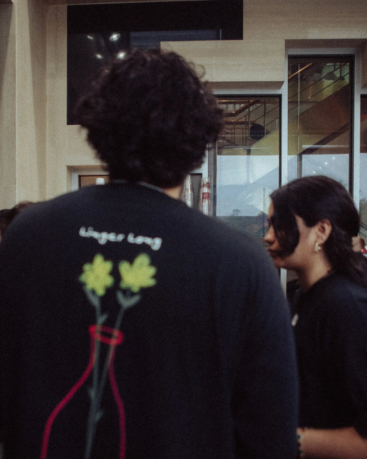 A person from behind wearing a black crewneck sweatshirt with a graphic of yellow flowers in a red vase and the text 'Linger Long' on the back, standing in an indoor setting.
